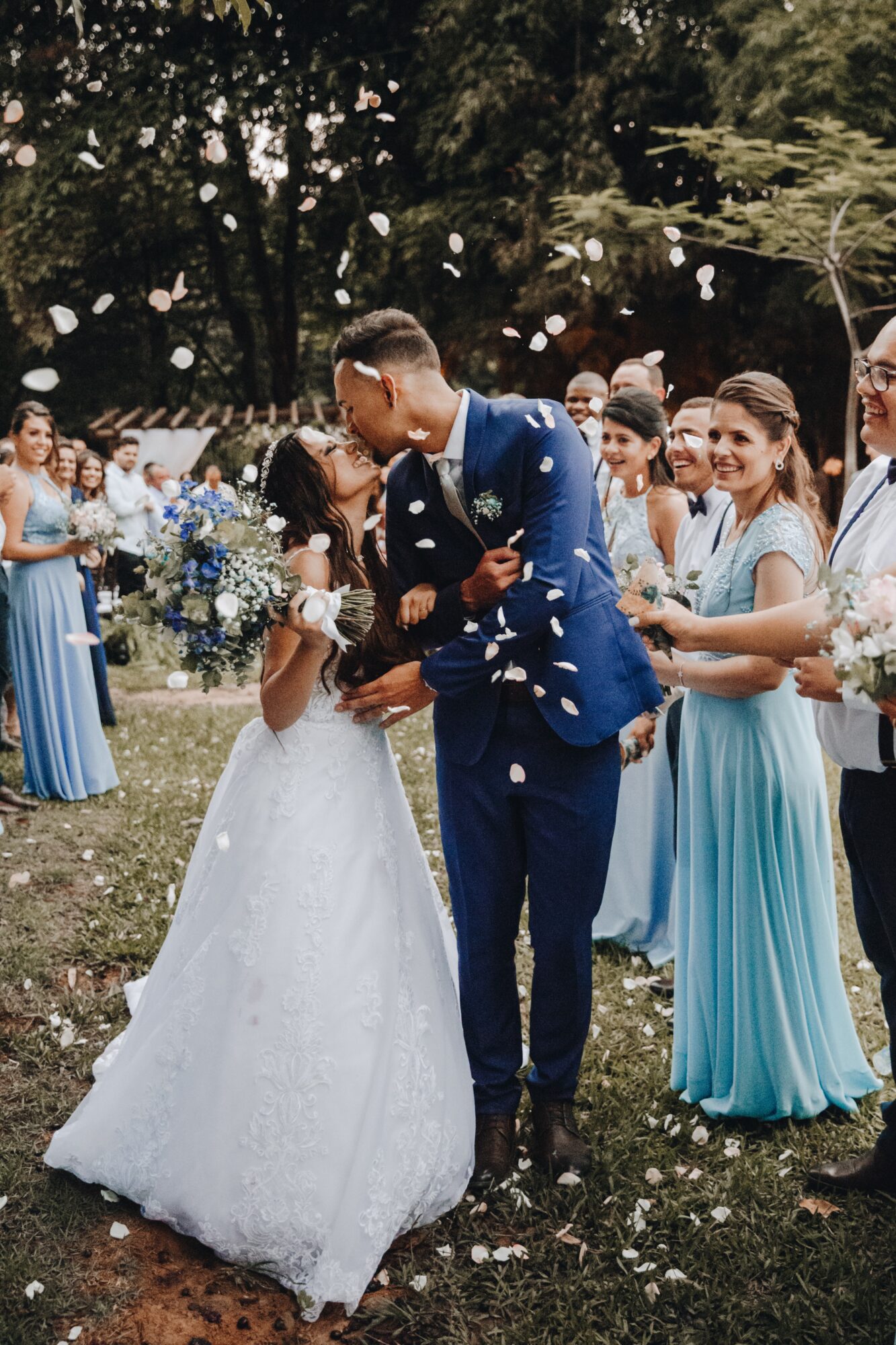 Couple de mariés embrassé sous une pluie de pétales entouré de demoiselles d'honneur en robes bleues.